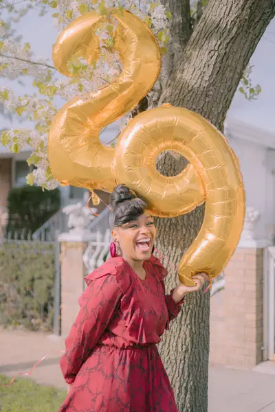 A laughing woman in a red patterned dress holds large gold 29 number balloons beside a blossoming tree in front of a brick house.