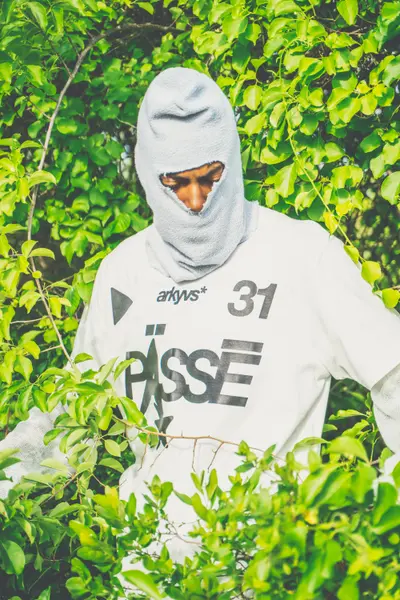 A masked person in a gray balaclava and white printed t-shirt stands partially hidden among bright green leaves and branches.