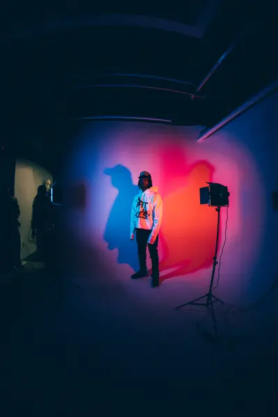 A man in a white hoodie and cap stands against a studio backdrop lit with blue and red lights, with a strobe on a stand beside him.