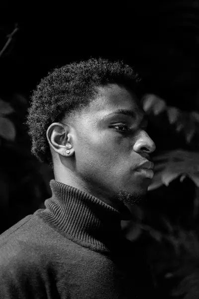 Black-and-white profile portrait of a young person with short curly hair and a stud earring, wearing a dark ribbed turtleneck against a shadowy background.