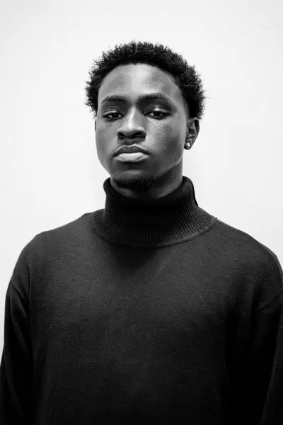 Young man with short curly hair and a goatee wearing a dark turtleneck sweater, posing against a light background in a black-and-white studio portrait.