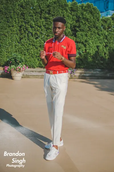A young man in a red polo shirt, white trousers, and white sneakers stands outdoors holding a pair of eyeglasses, with green hedges and flower planters behind him.