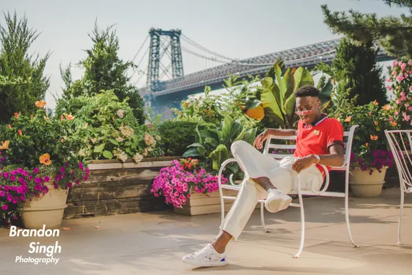 A young man in a red polo, white pants, and pink sunglasses sits on a white chair on a flower-filled rooftop terrace with a suspension bridge in the background.