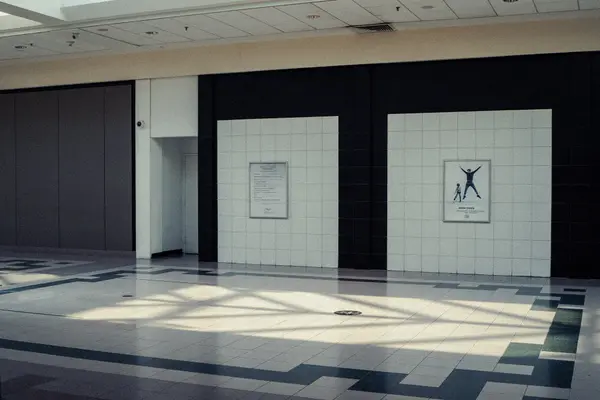 Empty mall interior with white and black tiled walls, geometric patterned floor, drop ceiling, and two framed posters reading WELCOME and NOW OPEN.