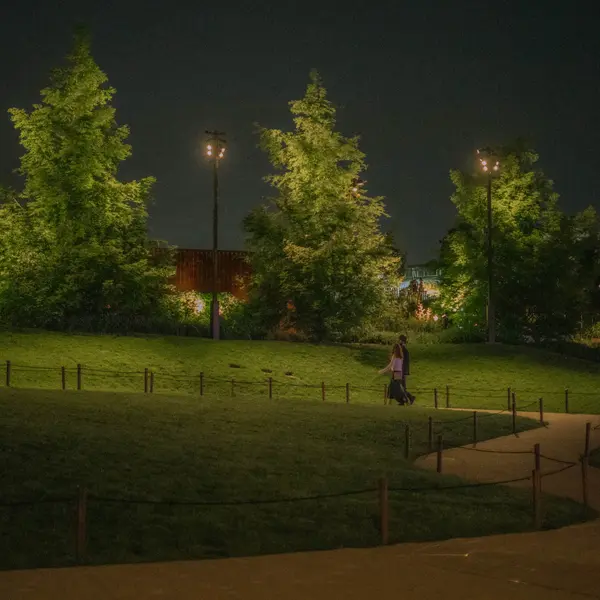 A couple walks along a path at night in a park, bordered by low posts and ropes, with illuminated trees and buildings in the background.
