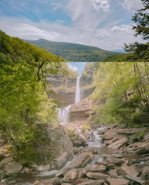 A tall waterfall cascades down a rocky cliff into a boulder-strewn stream, framed by green trees, with a panoramic view of forested mountains and cloudy sky above.