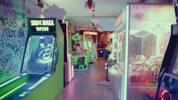 A dimly lit arcade corridor lined with glowing Skee-Ball and ticket machines, neon lights reflecting on the floor, and a graffiti-covered wall to the left.
