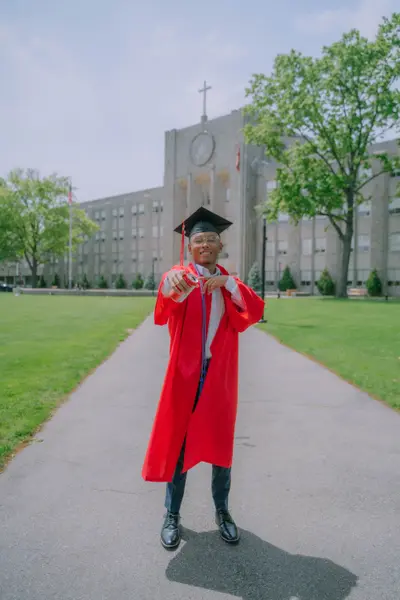 A smiling young man in a red graduation gown and cap holds a soda can, posing on a walkway in front of a large stone school building with a cross.