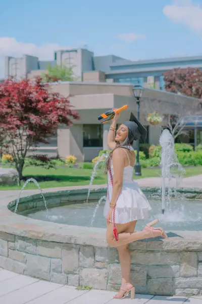 A woman in a white sundress and black graduation cap celebrates with a champagne bottle raised, posing on a stone fountain near a campus building.