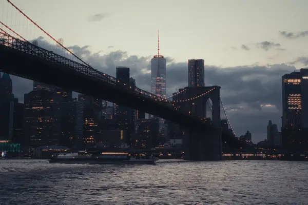 The illuminated Brooklyn Bridge spans the East River at dusk, with the Lower Manhattan skyline and One World Trade Center visible behind it.