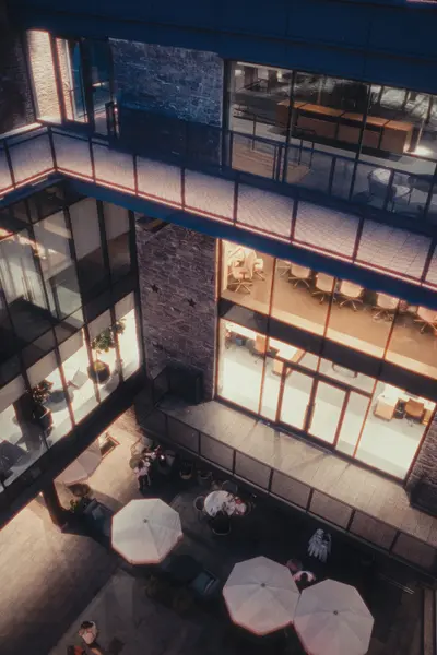 Aerial view of a multi-story modern atrium with glass-walled offices, exposed brick walls, warm interior lighting, and a ground-floor terrace with white umbrellas and seated people.